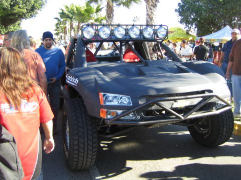 1990 Ford Bronco - black off-road trophy truck with a six-pod light bar and exposed tube front bumper, surrounded by spectators under palm trees.