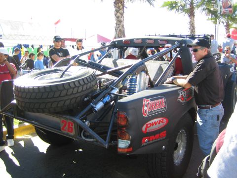 1990 Ford Bronco - Rear view of a dark off-road race truck, number 26, showing the spare tire, coilover shocks and exposed tube frame.