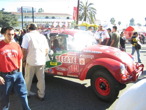 1990 Ford Bronco - red Volkswagen Beetle-based Baja Bug, number 1100, with Tecate livery, parked on a street among spectators.