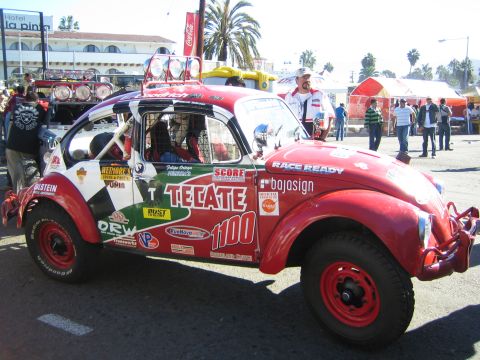 1990 Ford Bronco - Side view of a red Tecate-liveried Baja Bug, number 1100, with red wheels and a roof light bar in a busy lot.