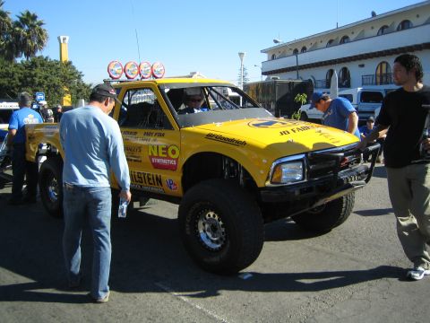 1990 Ford Bronco - yellow off-road race truck with a roof light bar, tube front bumper and sponsor decals, drawing spectators in a street.