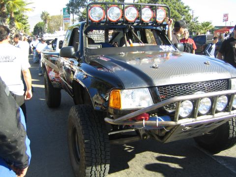 1990 Ford Bronco - black off-road race truck with a five-pod roof light bar and tube front bumper, surrounded by a crowd in a street.