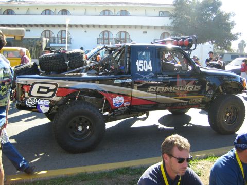 1990 Ford Bronco - black off-road race truck marked 1504 with Camburg livery and roof-mounted spare tires sits among spectators outside a white building.