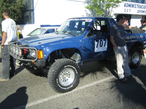 1990 Ford Bronco - blue lifted race pickup marked 707 with a front brush guard and winch is parked by a building as two men stand beside it.