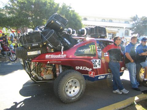 1990 Ford Bronco - dark red desert race buggy marked BC-4 piled high with gear bags and spare tires draws onlookers on a street.