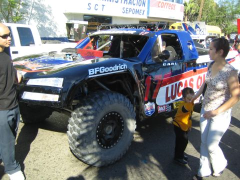 1990 Ford Bronco - black-and-blue trophy truck with Lucas Oil and BFGoodrich livery sits on a street as a woman and child walk past.