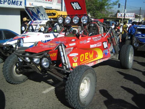 1990 Ford Bronco - red tube-frame desert buggy marked 104 with ORW livery and a bank of round driving lights sits at a busy staging area.