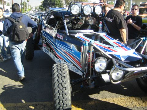 1990 Ford Bronco - black desert race buggy with red, white and blue star graphics, roof-mounted lights and King shocks sits among onlookers.