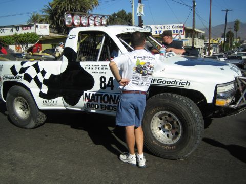 1990 Ford Bronco - white Chevrolet race pickup marked 84 with checkered-flag and National Rod Ends graphics is parked on a street as a man stands by it.