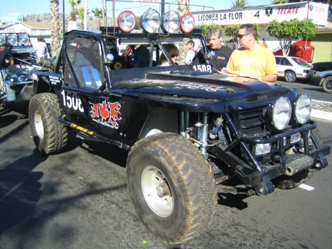 1990 Ford Bronco - black tube-frame race buggy marked 1508 with an ICE decal and roof-mounted lights is parked on a street with men beside it.