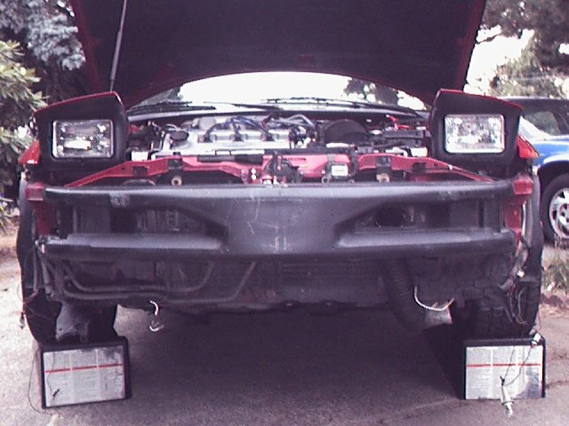 red car front end, hood up, engine exposed, bumper removed, sitting on blocks. blue car in background