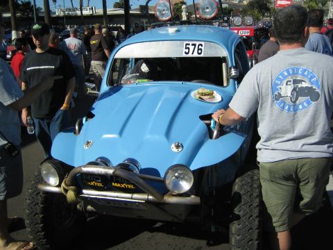 1990 Ford Bronco - light blue Baja Bug built from a VW Beetle, marked 576 with Maxtel decals and dual round lights, draws a crowd on a street.