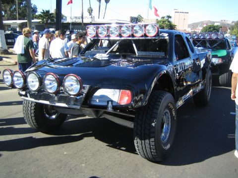 1990 Ford Bronco - black trophy truck with a retro coupe-style body, six round front lights and a roof light bar is parked at a staging area.