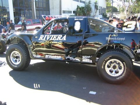 1990 Ford Bronco - glossy black race truck with Riviera Racing and number 3 livery on BFGoodrich tires is shown in side profile near a building.