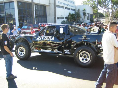 1990 Ford Bronco - black Riviera Racing race truck marked 3 sits in a street outside a Mexican Sector Comunicaciones building as people gather.
