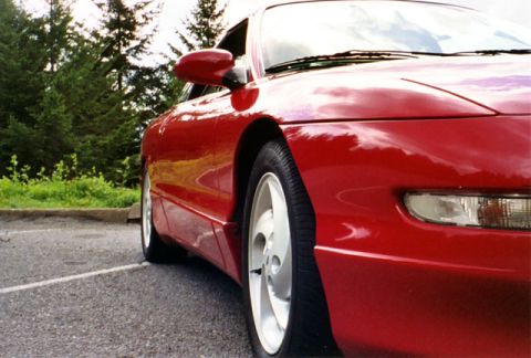 1996 Ford Probe - The front corner of a shiny red sports coupe is visible, parked outdoors with trees in the background.