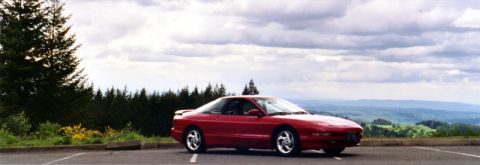 1996 Ford Probe - A red coupe is parked in a lot with a scenic view of rolling hills and a cloudy sky.