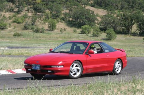 1996 Ford Probe - A red 1996 Ford Probe is captured in motion on a paved track, with a driver visible inside.