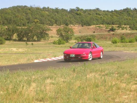 1996 Ford Probe - A red 1996 Ford Probe navigates a turn on a paved track surrounded by dry grass and rolling hills.