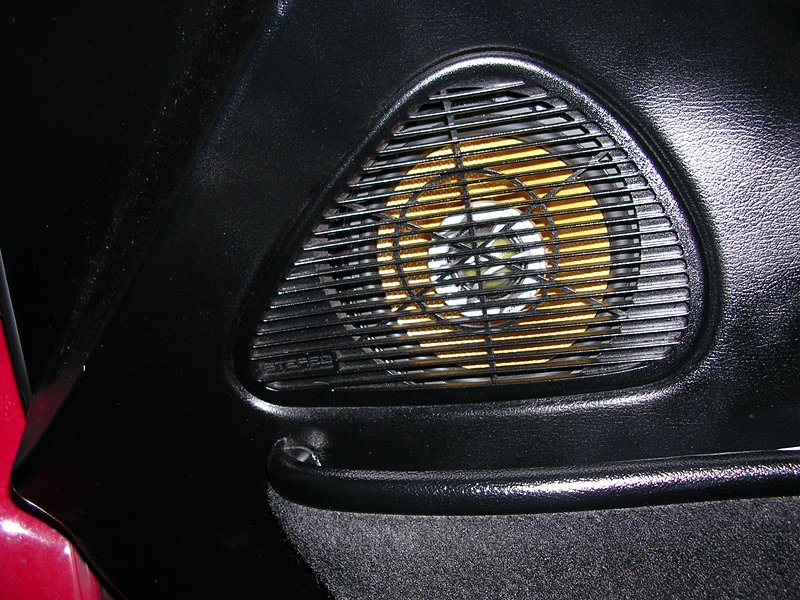 Black car door panel with a triangular speaker grille and yellow speaker cone visible
