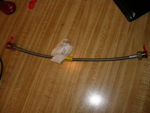 1996 Ford Probe - A braided metal hose with red caps on each end and a yellow tag with 'GOODRIDGE' printed on it lies on a wooden surface.