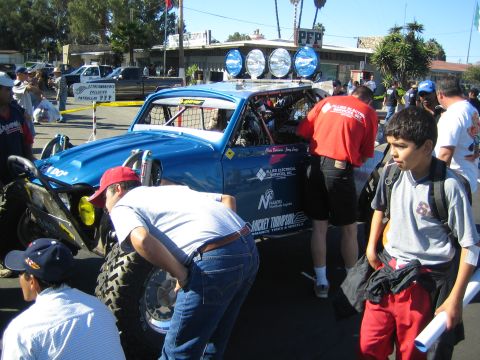 1990 Ford Bronco - blue Baja Bug built from a VW Beetle is surrounded by crew and spectators as people work on it at a street staging area.