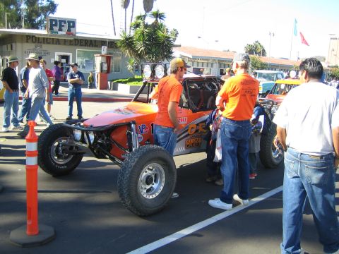 1990 Ford Bronco - orange tube-frame desert race buggy is attended by a crew in orange shirts on a street outside a Policia Federal building.
