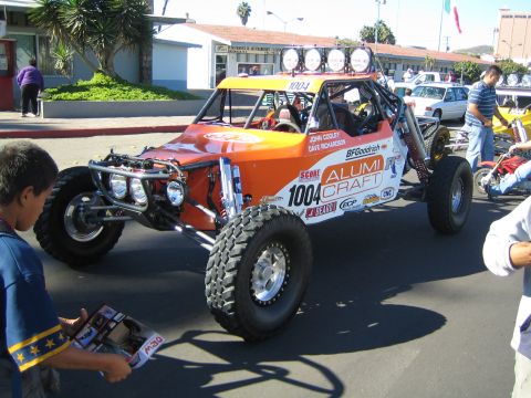 1990 Ford Bronco - orange Alumi Craft desert race buggy marked 1004 with a bank of roof lights is parked on a street as people gather.