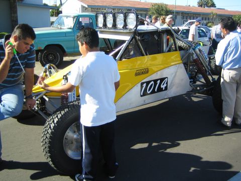 1990 Ford Bronco - yellow-and-white tube-frame race buggy marked 1014 is worked on by people at a staging lot, with a vintage pickup parked behind.