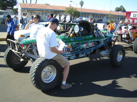 1990 Ford Bronco - green single-seat off-road race buggy with an exposed tube chassis and mid-mounted engine sits curbside while two men inspect it.