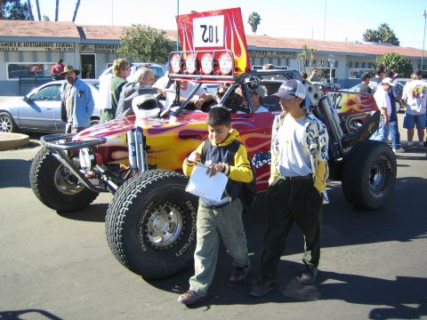 1990 Ford Bronco - off-road race buggy painted with red-and-yellow flames and a number 102 roof banner draws onlookers, including two boys in front.