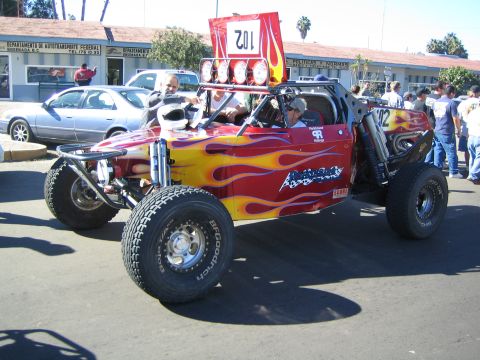 1990 Ford Bronco - red off-road race buggy with yellow flame graphics and a 102 roof banner rolls along a street past parked cars and spectators.