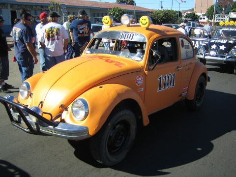 1990 Ford Bronco - orange Volkswagen Beetle built as a Baja race bug, numbered 1101 with roof-mounted lights and smiley-face decals, parked among a crowd.