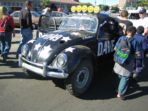 1990 Ford Bronco - black Volkswagen Beetle Baja race bug marked with white stars and DAVE lettering and roof-mounted smiley lights stands amid spectators.