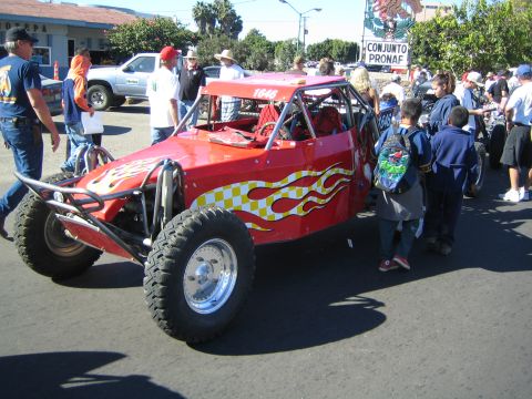 1990 Ford Bronco - red open-cockpit off-road buggy with yellow flame and checkered graphics is surrounded by spectators on a sunny street.