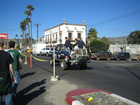 1990 Ford Bronco - blue off-road truck loaded with several riders drives away through a street intersection in front of a historic building and palm trees.