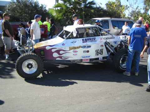 1990 Ford Bronco - white off-road race buggy with purple flames, Yokohama and Savage sponsor decals and number 1649 stands among a crowd of onlookers.