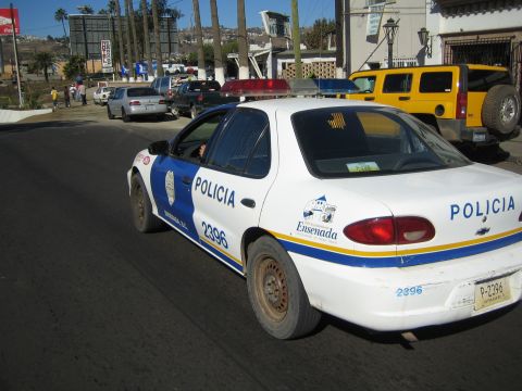 1990 Ford Bronco - white Chevrolet sedan painted as an Ensenada police car, numbered 2396, drives down a street with a yellow SUV parked in the background.