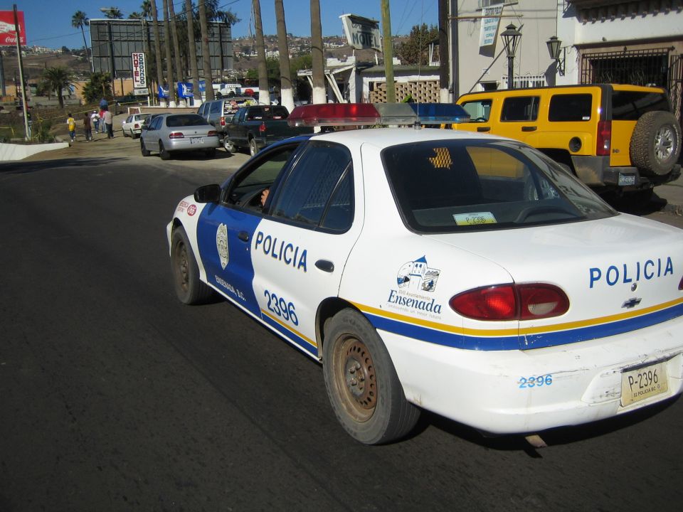 White Chevrolet sedan marked POLICIA Ensenada number 2396 on a street with a yellow SUV behind