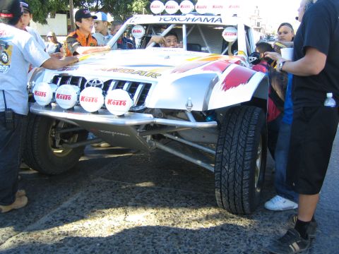 1990 Ford Bronco - front of a silver off-road race buggy fitted with a row of Koito driving lights and Yokohama branding is surrounded by people on a cobble s…