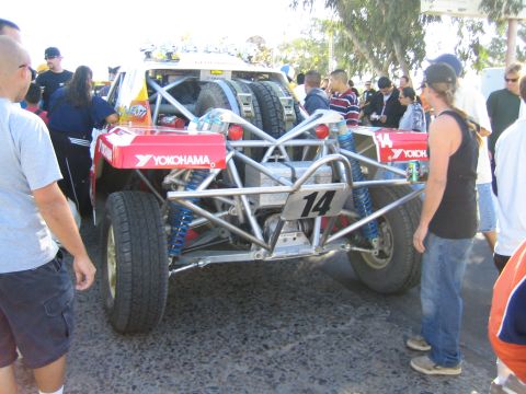 1990 Ford Bronco - rear of a red-and-white off-road race buggy numbered 14, with Yokohama decals, coilover shocks and twin spare tires, faces a crowd.