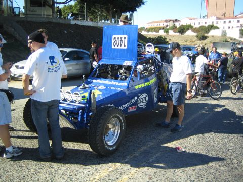 1990 Ford Bronco - blue off-road race buggy numbered 1405 with sponsor decals is inspected by crew while spectators and bicycles stand nearby.