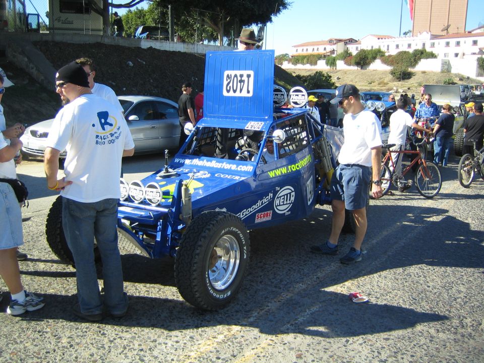 Blue off-road race buggy number 1405 with sponsor decals and bicycles nearby on a paved lot
