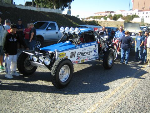 1990 Ford Bronco - blue-and-white tube-frame off-road buggy numbered 1614 with BFGoodrich decals and roof lights is surrounded by a crowd on a paved lot.