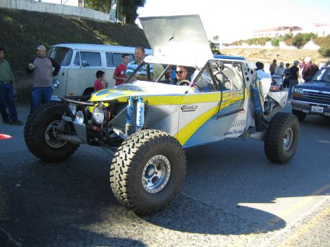 1990 Ford Bronco - silver-and-yellow off-road race buggy with its rear hood raised and Goodyear branding sits on a street as spectators and an old VW van look…