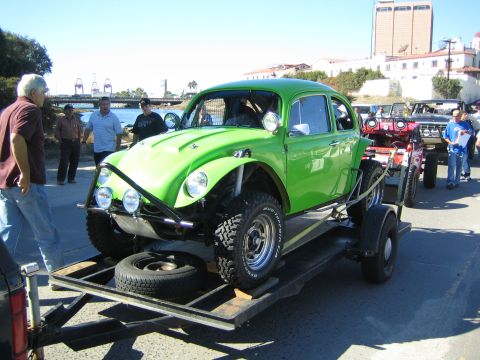 1990 Ford Bronco - bright green Volkswagen Beetle Baja race bug sits strapped on an open trailer beside a waterfront with port cranes in the distance.
