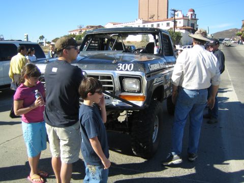 1990 Ford Bronco - black Ford off-road race truck numbered 300 stands on a street as a crowd including children gathers around its front fender.
