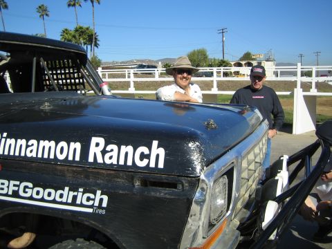 1990 Ford Bronco - Two men stand behind the hood of a black Ford race truck lettered Cinnamon Ranch and BFGoodrich, with a white fence and palms beyond.
