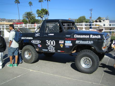 1990 Ford Bronco - side view of a black Ford race pickup numbered 300, lettered Cinnamon Ranch and BFGoodrich, parked by a white fence with palms and onlooker…