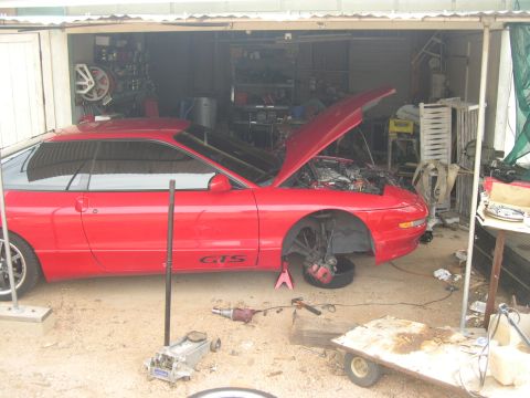 1997 Ford Probe - A red coupe, identified as a 1990 model by its body style, is undergoing repairs in a garage with its hood and front wheels removed.
